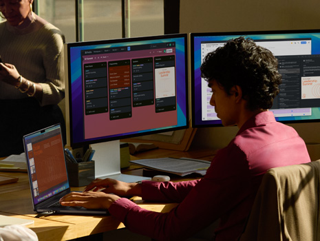 A woman sitting at a table in a highrise building. Behind her is a large window revealing other buildings. In front of her is an open MacBook.