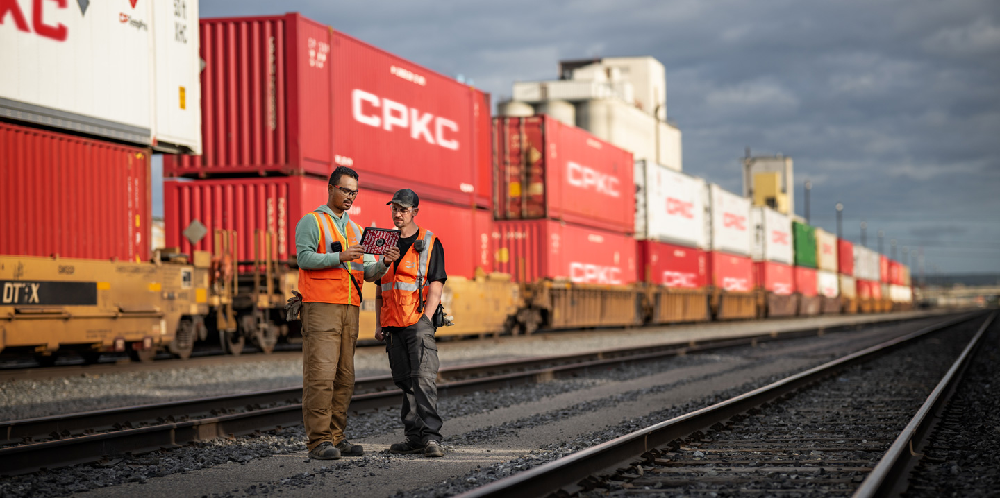 Hai công nhân đường sắt đang tham khảo thông tin trên iPad khi đứng trong sân ga, bên cạnh một đoàn tàu chở container đang đứng yên