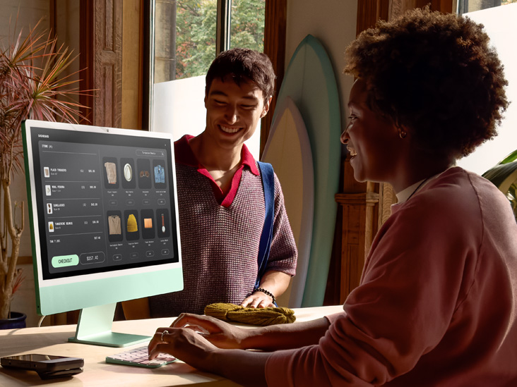 Hai người trong một căn phòng đủ ánh sáng, một người đang kiểm tra đơn đặt hàng trên một chiếc iMac M4 màu xanh lá cây.