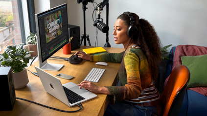 A woman in her home working at her desk with MacBook Pro connected to two external displays, wearing headphones and speaking into a microphone